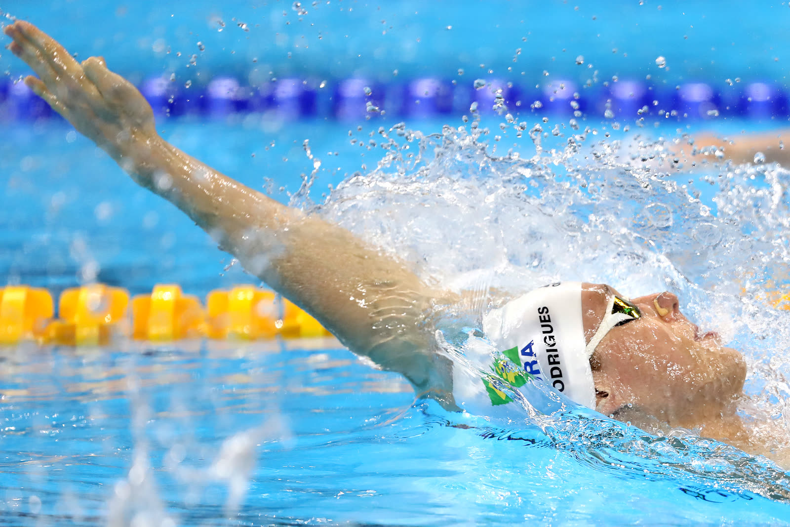Henrique Rodrigues racing backstroke in competition, arm sweeping overhead as water flies off the stroke